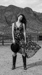 fashion model in flowing floral dress and cowboy boots standing in desert landscape with mountains holding black hat