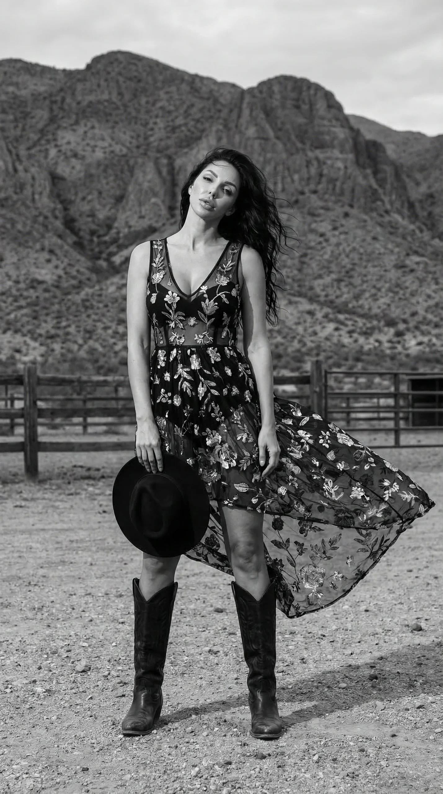 fashion model in flowing floral dress and cowboy boots standing in desert landscape with mountains holding black hat